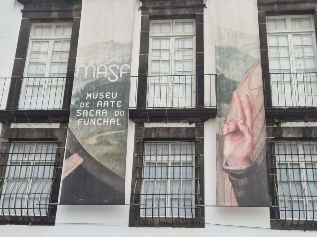 Voorgevel van het Museu de Arte Sacra do Funchal met twee verticale doeken: links de naam van het museum, rechts een detail van een religieus schilderij met een hand in zegenend gebaar"